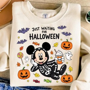 May include: Cream-colored sweatshirt with a Halloween-themed graphic. The design features Mickey Mouse in a skeleton costume, pumpkins, bats, candy, and the text "JUST WAITING FOR HALLOWEEN".