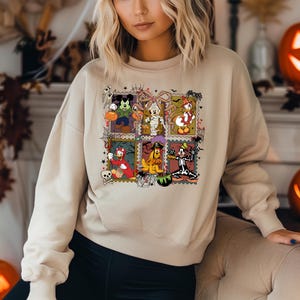 May include: Beige sweatshirt featuring a colorful Halloween-themed graphic. The graphic is composed of postage stamp-style images of Disney characters dressed in Halloween costumes, including Mickey Mouse, Donald Duck, and Goofy.