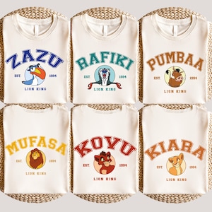May include: Six off-white t-shirts featuring Lion King characters: Zazu, Rafiki, Pumbaa, Mufasa, Kovu, and Kiara. Each shirt has the character's name in a collegiate font, the text "EST. 1994", and a character illustration. The shirts are displayed on a woven surface.