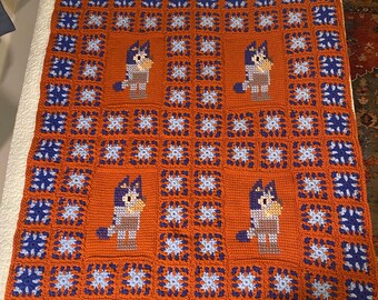 May include: Handmade crochet blanket featuring a repeating pattern of squares in shades of orange, blue, and light blue. The blanket showcases four central squares with a cartoon dog design. The blanket is a cozy, handmade item.