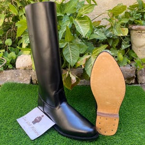 Op de afbeelding: Een hoge, zwarte leren rijlaars met een gladde afwerking. De lichtbruine zool heeft een textuur. Een klein papiertje met tekst ligt naast de laars. De laars staat op een groene ondergrond, met planten op de achtergrond.