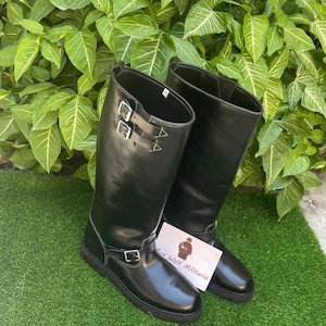 Puede incluir: Un par de botas altas de cuero negro con hebillas y correas plateadas. Las botas son hasta la rodilla con un acabado brillante, sobre un fondo de follaje verde y césped artificial. Se ve una tarjeta con el texto "Wolf Militaria".