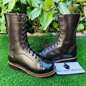 Könnte beinhalten: Ein Paar schwarze Leder-Kampfstiefel mit Schnürung. Die Stiefel haben eine glänzende Oberfläche und eine braune Sohle. Die Stiefel werden auf grünem Gras präsentiert. Eine weiße Karte mit dem Text "Zee Wolf Militaria" ist ebenfalls vorhanden.