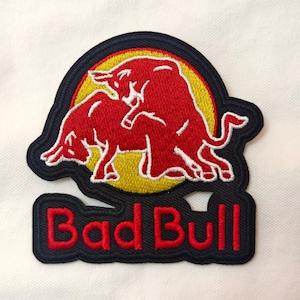 Könnte beinhalten: Gestickter Aufnäher mit zwei roten Bullen auf gelbem Grund, umrandet von Marineblau. Der Text "Bad Bull" ist rot. Dieser Aufnäher ist für Kleidung oder Accessoires gedacht und setzt ein klares Zeichen.
