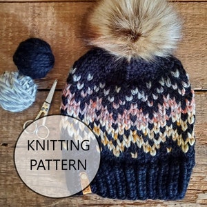 Op de afbeelding: Breipatroon voor een zwart-witte gebreide muts met een bruine nep bont pompon. De muts heeft een geometrisch ontwerp met roze, witte en gele accenten.