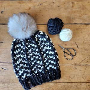 May include: Black and white knit hat with a white pom pom. The hat is sitting on a wooden surface with two balls of yarn and a pair of scissors.