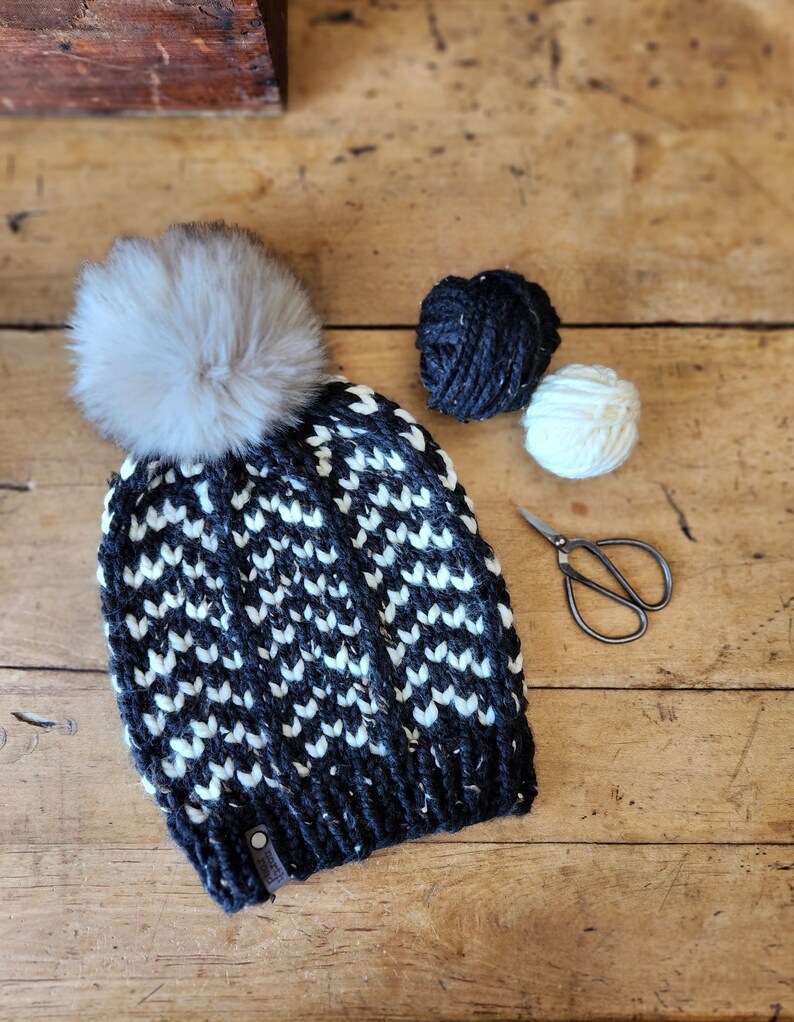 May include: Black and white knit hat with a white pom pom. The hat is sitting on a wooden surface with two balls of yarn and a pair of scissors.