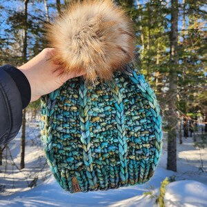 May include: A teal, blue, and green knitted beanie with a brown faux fur pom pom. The beanie has a brown leather tag with the text "NORTH" on it.