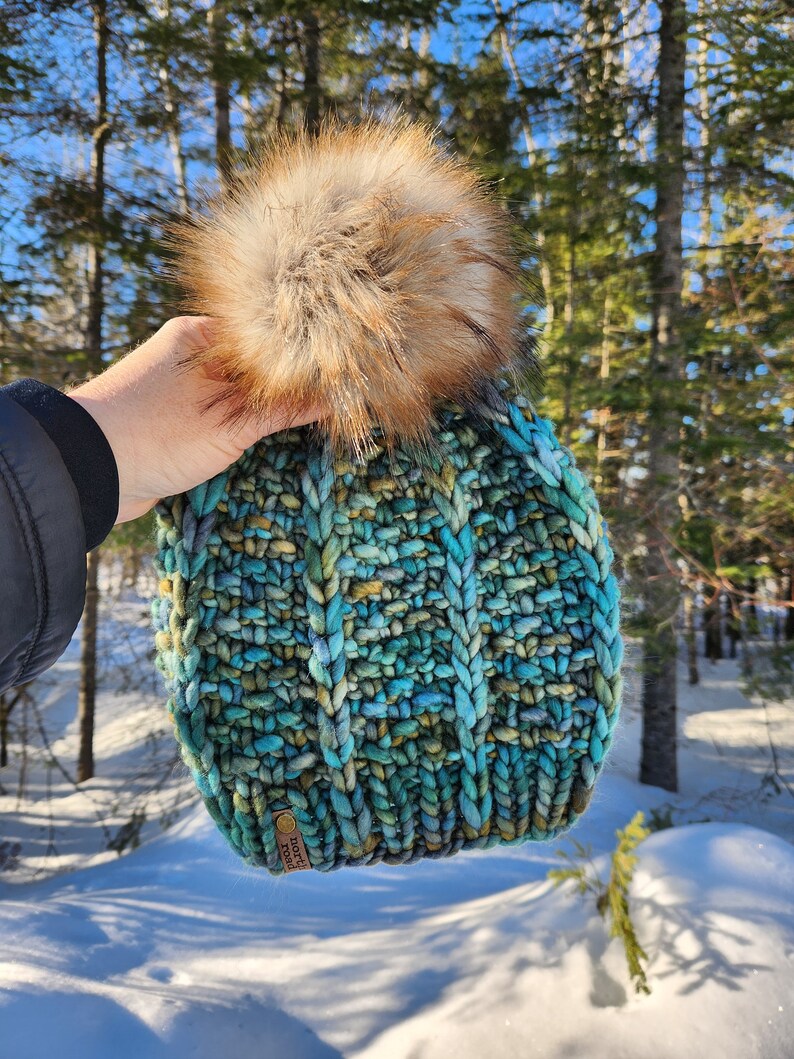May include: A teal, blue, and green knitted beanie with a brown faux fur pom pom. The beanie has a brown leather tag with the text "NORTH" on it.