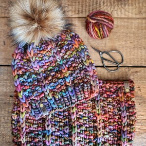 May include: A colorful knitted hat and matching scarf with a brown faux fur pom pom. The hat and scarf are made with a chunky yarn in a variety of colors, including purple, blue, green, red, and orange.