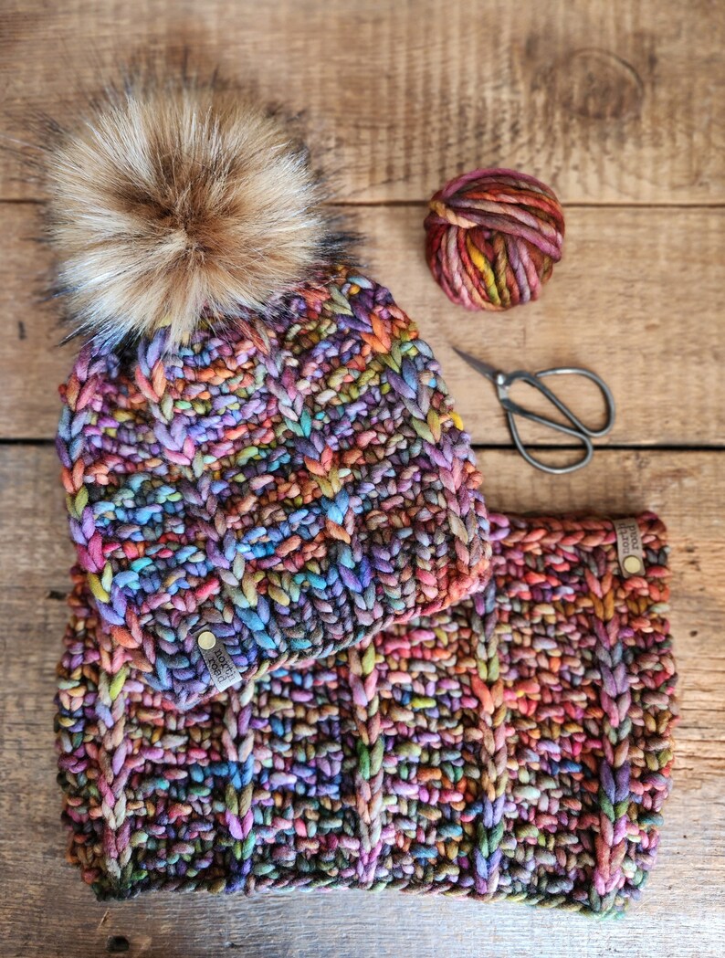 May include: A colorful knitted hat and matching scarf with a brown faux fur pom pom. The hat and scarf are made with a chunky yarn in a variety of colors, including purple, blue, green, red, and orange.