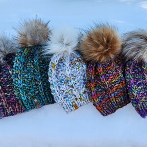 May include: Five colorful knitted hats with faux fur pom poms. The hats are in shades of purple, blue, white, red, and green.