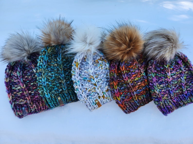 May include: Five colorful knitted hats with faux fur pom poms. The hats are in shades of purple, blue, white, red, and green.