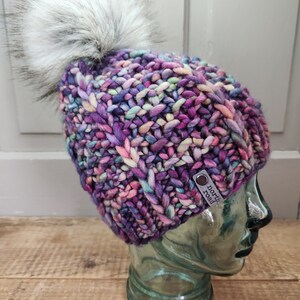 May include: A colorful knitted beanie with a fluffy white pom pom. The beanie has a purple, pink, blue, and green color scheme. The beanie has a leather tag that says "North Road".