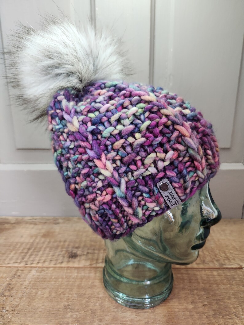 May include: A colorful knitted beanie with a fluffy white pom pom. The beanie has a purple, pink, blue, and green color scheme. The beanie has a leather tag that says "North Road".