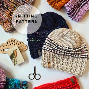 May include: A collection of chunky knit hats and headbands in various colors including navy, cream, orange, red, and yellow. A wooden knitting gauge, a small tin with a bird illustration, and black scissors are arranged among the knit items.