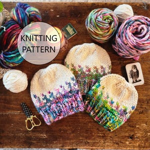 Op de afbeelding: Drie kleurrijke gebreide mutsen met een witte achtergrond en een veelkleurig patroon. De mutsen liggen op een houten tafel met garenbollen en breiaccessoires. De tekst "Knitting Pattern" is zichtbaar in de afbeelding.