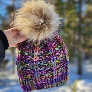 May include: A colorful knitted beanie with a fluffy pom pom on top. The beanie has a brown leather tag with the text "north" and "fork" on it.