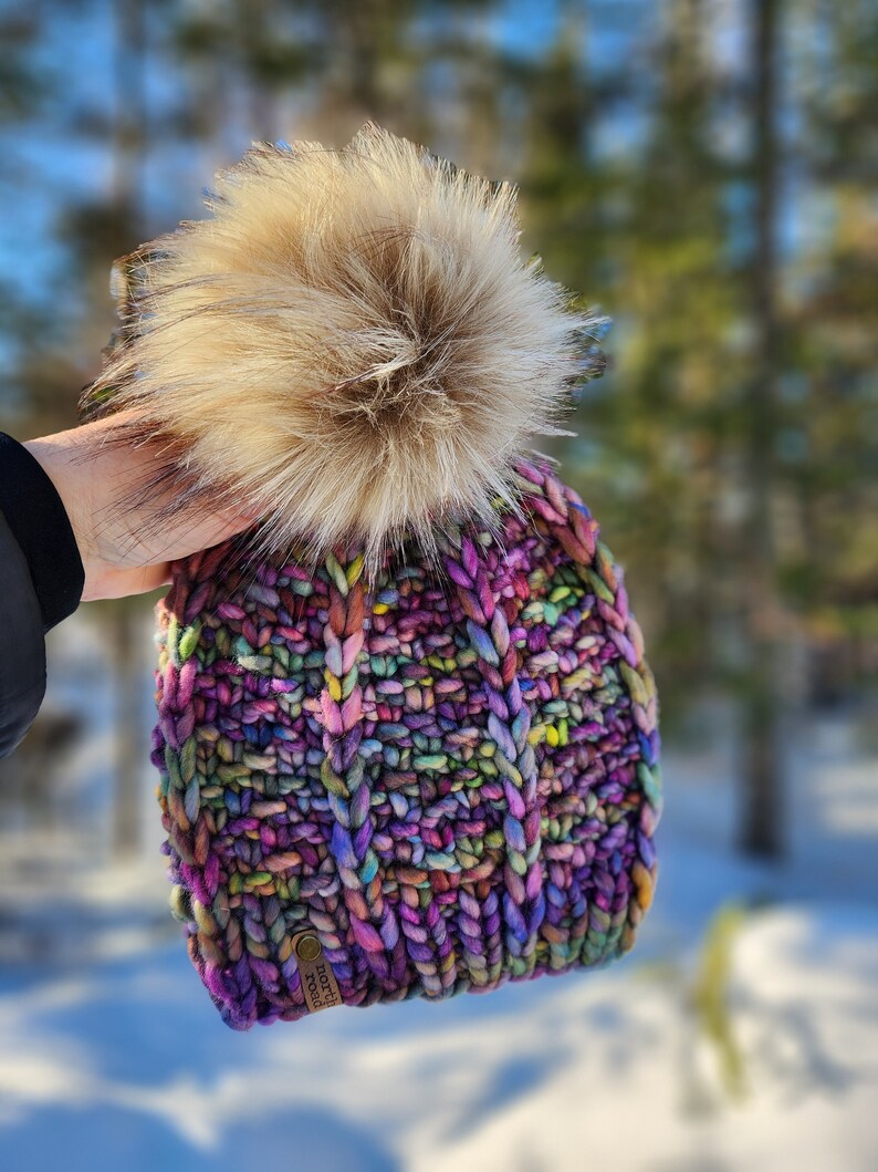 May include: A colorful knitted beanie with a fluffy pom pom on top. The beanie has a brown leather tag with the text "north" and "fork" on it.