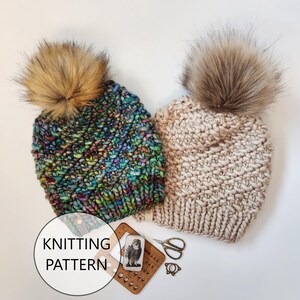 May include: Two knitted hats with faux fur pom poms. One hat is a multi-colored yarn with a blue, green, and purple color scheme. The other hat is a solid beige color. The hats are laying on a white surface with knitting supplies.