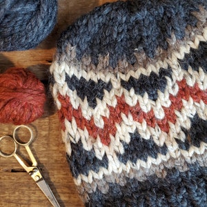 May include: A close-up of a knitted hat with a gray, white, and orange chevron pattern. The hat is made with thick yarn and has a fuzzy texture. There are also balls of yarn and a pair of scissors in the image.