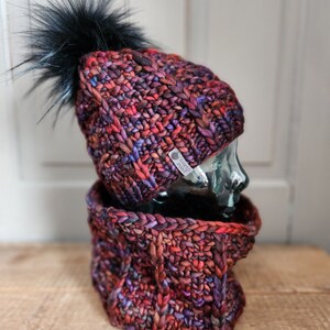 May include: A red, purple, and black knitted hat with a black pom-pom and a matching neck warmer. The hat has a small tag that says "Knot Too Knotty".