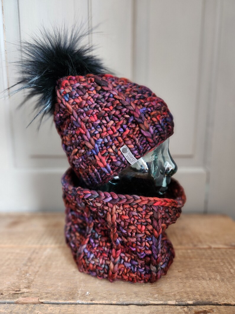 May include: A red, purple, and black knitted hat with a black pom-pom and a matching neck warmer. The hat has a small tag that says "Knot Too Knotty".