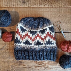 May include: A gray, white, and orange knitted hat with a geometric pattern. The hat is surrounded by balls of yarn in various colors, including gray, white, orange, and brown. There are also knitting needles and a pair of scissors.