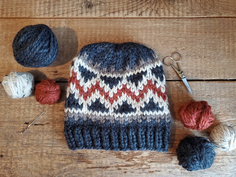 May include: A gray, white, and orange knitted hat with a geometric pattern. The hat is surrounded by balls of yarn in various colors, including gray, white, orange, and brown. There are also knitting needles and a pair of scissors.