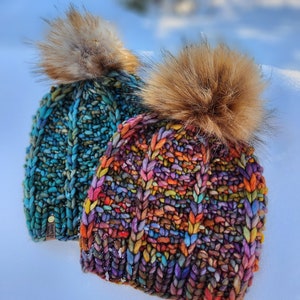 May include: Two colorful knitted hats with faux fur pom poms. The hats are made with a chunky yarn and have a unique, textured stitch pattern. The hats are sitting on a white surface.