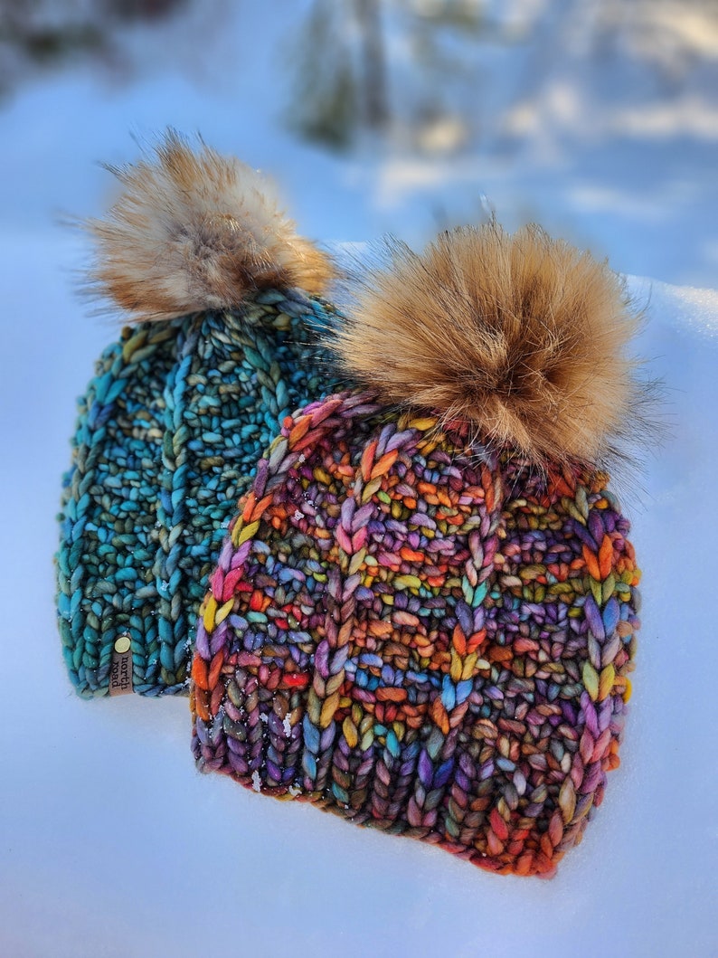 May include: Two colorful knitted hats with faux fur pom poms. The hats are made with a chunky yarn and have a unique, textured stitch pattern. The hats are sitting on a white surface.