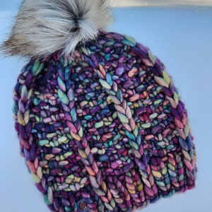May include: A colorful knitted beanie with a fluffy white pom pom. The beanie has a cable knit pattern and is made with a variety of colors, including purple, pink, blue, and green. The beanie has a small tag that says "north road" on it.