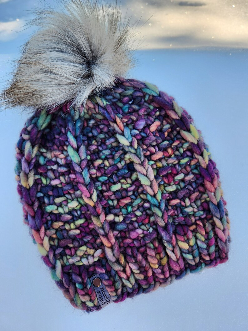 May include: A colorful knitted beanie with a fluffy white pom pom. The beanie has a cable knit pattern and is made with a variety of colors, including purple, pink, blue, and green. The beanie has a small tag that says "north road" on it.