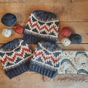 May include: Three knitted hats with a geometric pattern in shades of gray, red, and white. The hats are arranged on a wooden surface with yarn balls and other craft supplies.