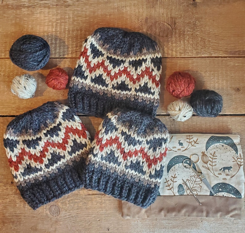 May include: Three knitted hats with a geometric pattern in shades of gray, red, and white. The hats are arranged on a wooden surface with yarn balls and other craft supplies.