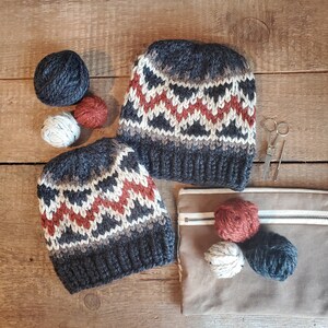 May include: Two black and white knit hats with a red and white geometric pattern. The hats are on a wooden surface with yarn balls and a small pair of scissors.