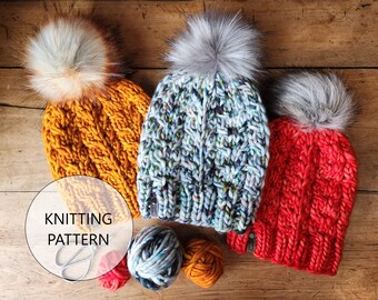 May include: Three knitted beanies with faux fur pom-poms in mustard yellow, speckled blue, and red. The beanies are made of thick yarn with a cable knit design. The image also shows balls of yarn and the text "KNITTING PATTERN".