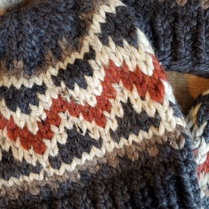 May include: A close-up of a knitted hat with a black, white, brown, and red geometric pattern. The hat is made of wool and has a soft, fuzzy texture.