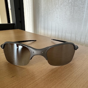 Pode incluir: Um par de &oacute;culos de sol com arma&ccedil;&atilde;o cinza fosco e lentes tingidas de marrom. Os &oacute;culos de sol est&atilde;o sobre uma superf&iacute;cie de madeira marrom clara. As lentes t&ecirc;m uma ligeira curvatura.