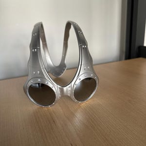 May include: Silver sunglasses with a distinctive, wraparound style. The lenses are dark and reflective, and the frame has small, decorative rivets. The bridge curves upwards, linking the two lenses. The frame is made of a metallic material.