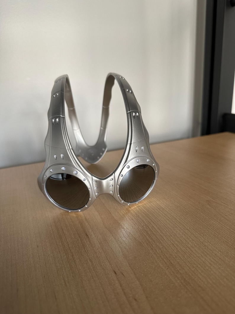 May include: Silver sunglasses with a distinctive, wraparound style. The lenses are dark and reflective, and the frame has small, decorative rivets. The bridge curves upwards, linking the two lenses. The frame is made of a metallic material.