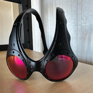 May include: Black sunglasses with a wraparound design and red lenses. The sunglasses have a thick frame with visible rivets and a curved temple. The lenses are round and reflect a pink hue. The sunglasses are resting on a wooden surface.