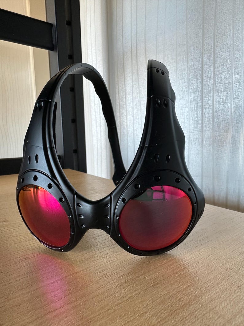 May include: Black sunglasses with a wraparound design and red lenses. The sunglasses have a thick frame with visible rivets and a curved temple. The lenses are round and reflect a pink hue. The sunglasses are resting on a wooden surface.