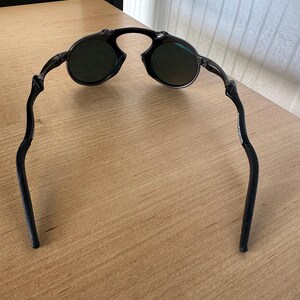 Pu&ograve; includere: Occhiali da sole neri con lenti rotonde e montatura curva. Gli occhiali da sole sono appoggiati su una superficie di legno marrone chiaro. Le lenti sembrano essere di colore verde scuro.