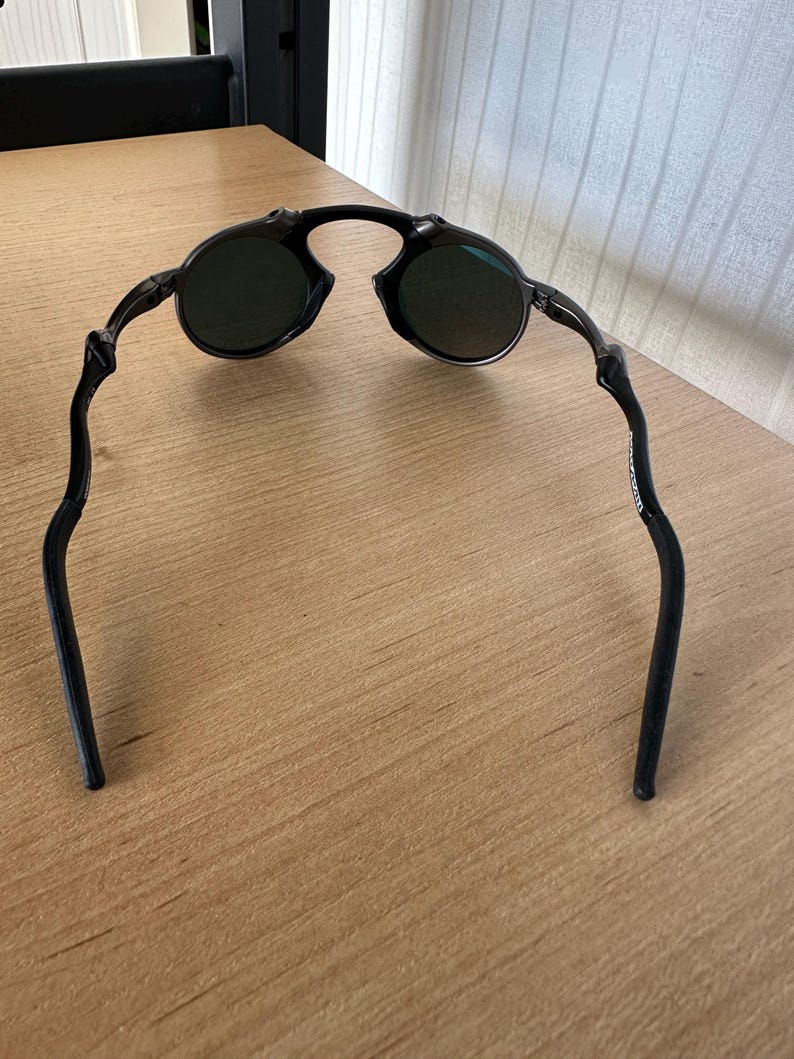 Pu&ograve; includere: Occhiali da sole neri con lenti rotonde e montatura curva. Gli occhiali da sole sono appoggiati su una superficie di legno marrone chiaro. Le lenti sembrano essere di colore verde scuro.