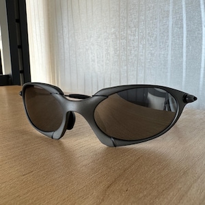 May include: Dark gray sunglasses with a curved frame and brown tinted lenses. The sunglasses are resting on a light brown wooden surface. The lenses reflect the surrounding environment.