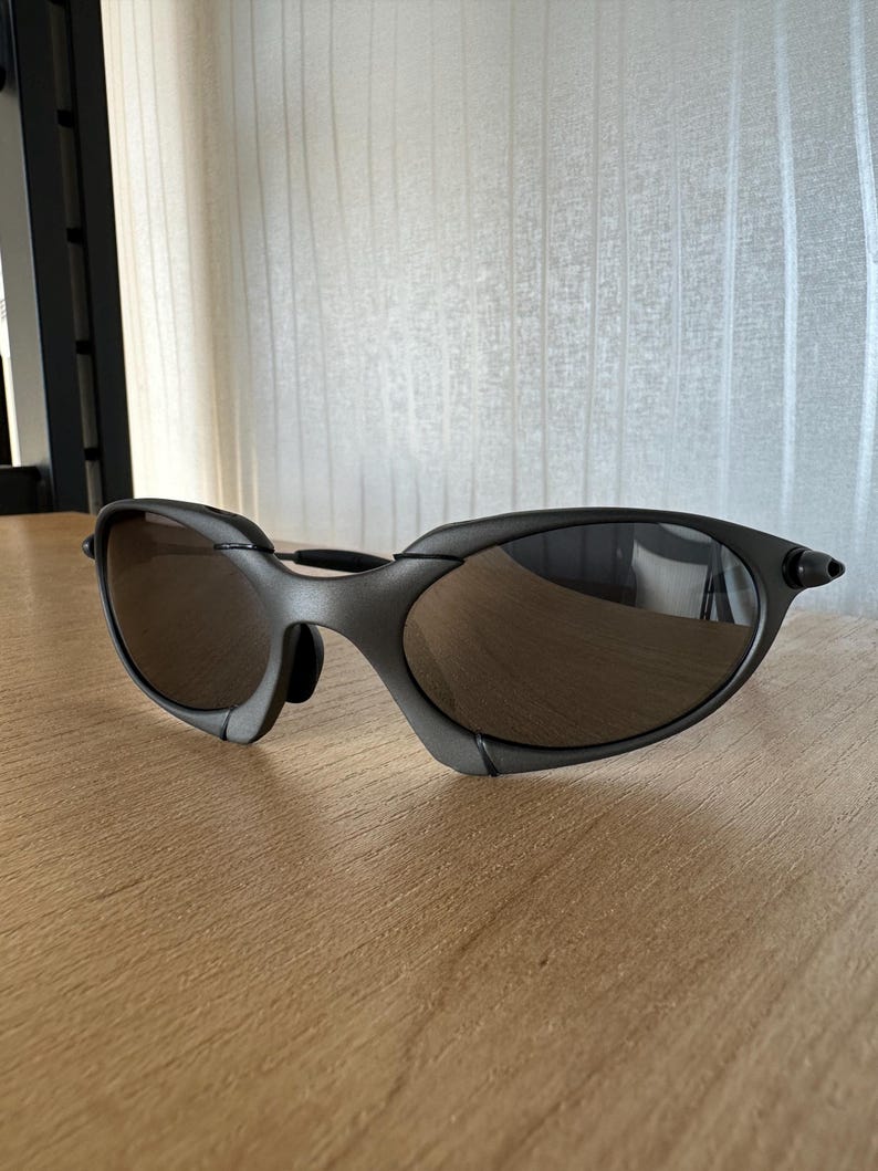 May include: Dark gray sunglasses with a curved frame and brown tinted lenses. The sunglasses are resting on a light brown wooden surface. The lenses reflect the surrounding environment.