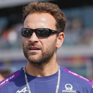 May include: A man wearing black sunglasses and a blue shirt with white and purple accents. The shirt features the Alpine and Pramac logos. The man has short brown hair and a beard. The background is blurred.