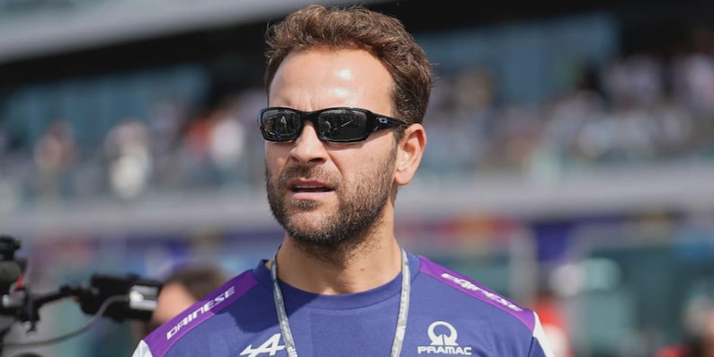 May include: A man wearing black sunglasses and a blue shirt with white and purple accents. The shirt features the Alpine and Pramac logos. The man has short brown hair and a beard. The background is blurred.