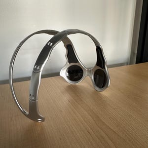 May include: Silver sunglasses with dark lenses. The sunglasses have a distinctive wraparound design with a curved frame and a bridge connecting the lenses. The lenses are oval-shaped and the frame is a metallic silver colour.